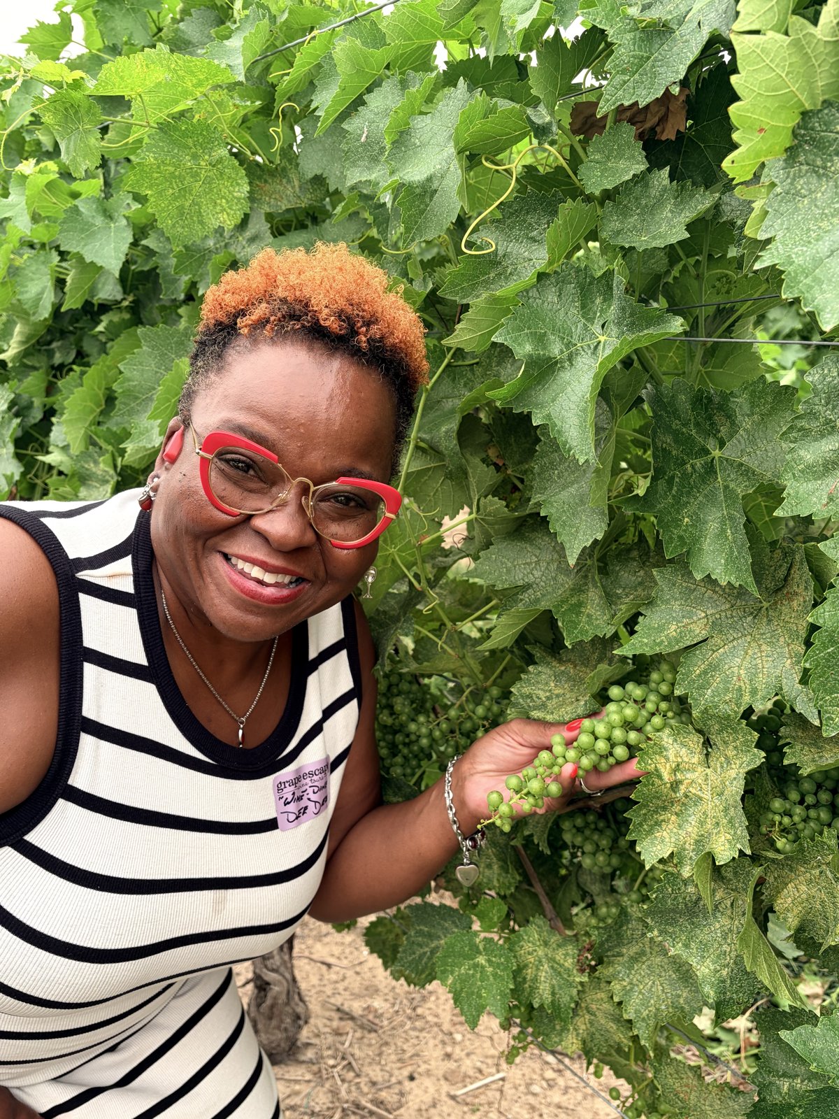 DeeDee Johnson smiling in a vineyard, holding a cluster of young grapes on the vine