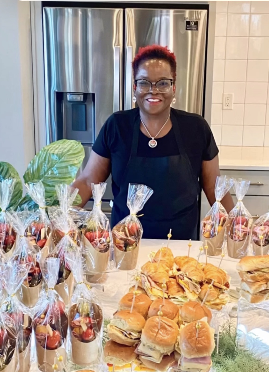 DeeDee smiling behind a catering spread of wrapped fruit skewers and croissant sliders, ready to serve guests